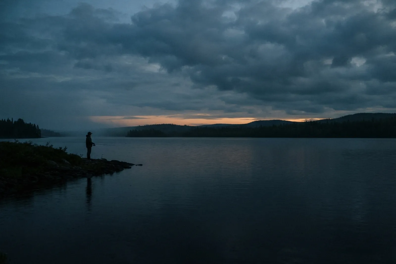 Lone angler on a dark lake shore at dusk under heavy cloud, the last orange light low on the horizon